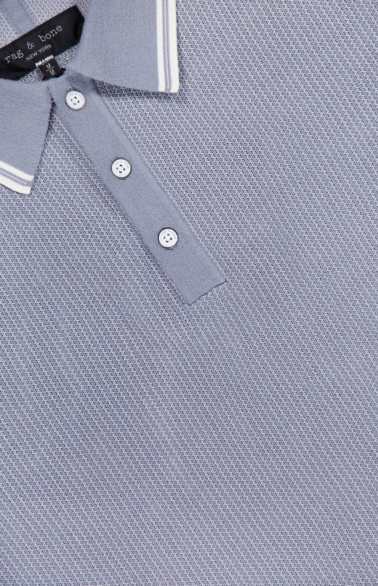 A close-up of the RAG & BONE Harvey Tipped Polo Shirt in light grey shows white buttons and a ribbed collar with two thin white stripes, plus the partially visible shirt tag at the neckline.