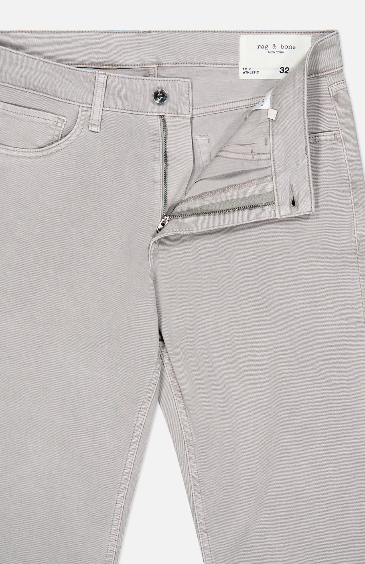 The Fit 3 Aero Stretch Pant in light grey, featuring a partially open zipper and button fly, displays the waistband label with the RAG & BONE brand and size 32. Crafted from comfortable denim with Aero Stretch technology, these pants are laid flat against a plain background.