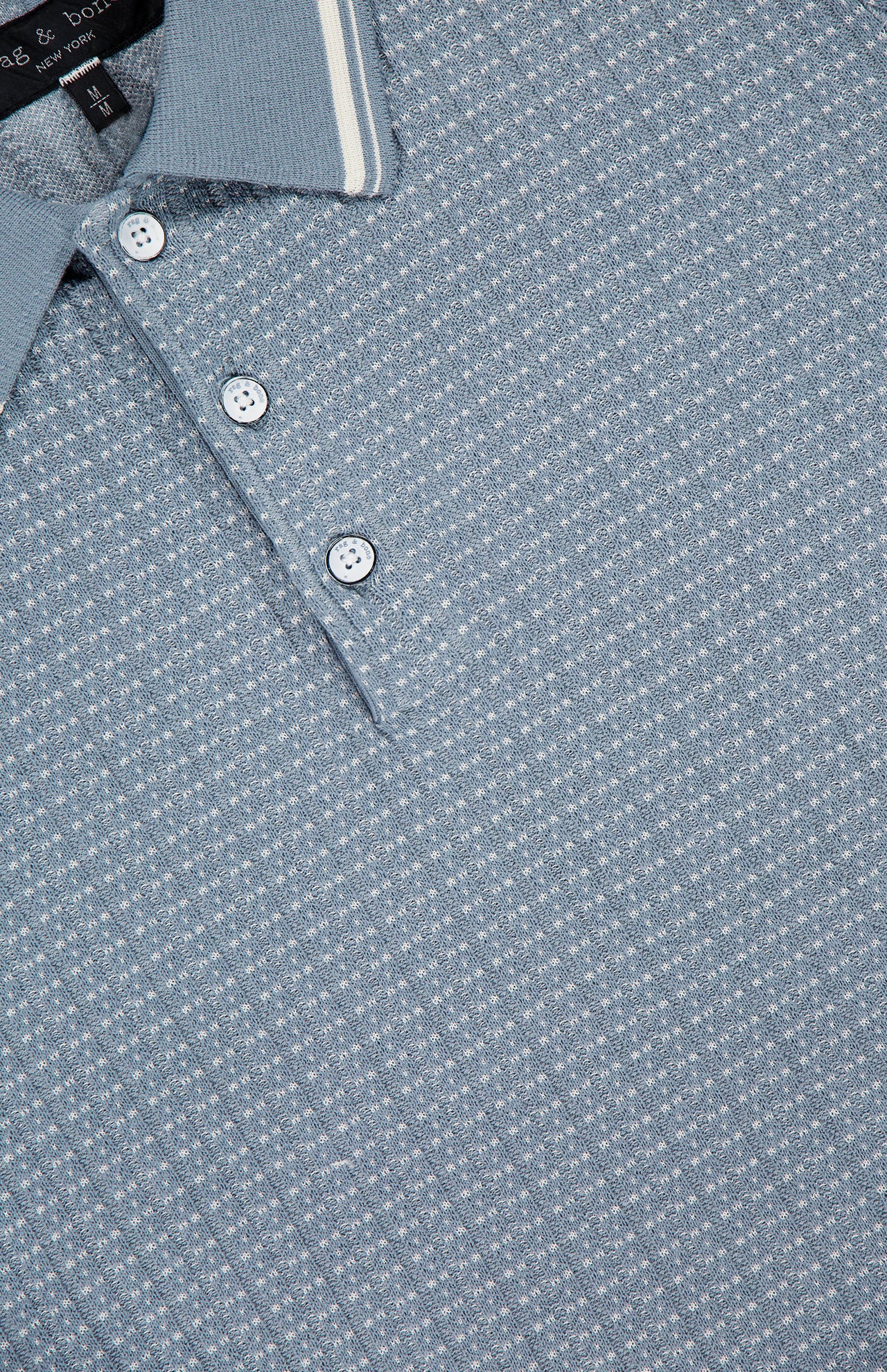 Close-up of the RAG & BONE Harvey Rib Polo Shirt, a blue-grey sweater with a vintage vibe, featuring a light blue pattern, white stripe on the collar, three white buttons, and a subtle geometric texture.