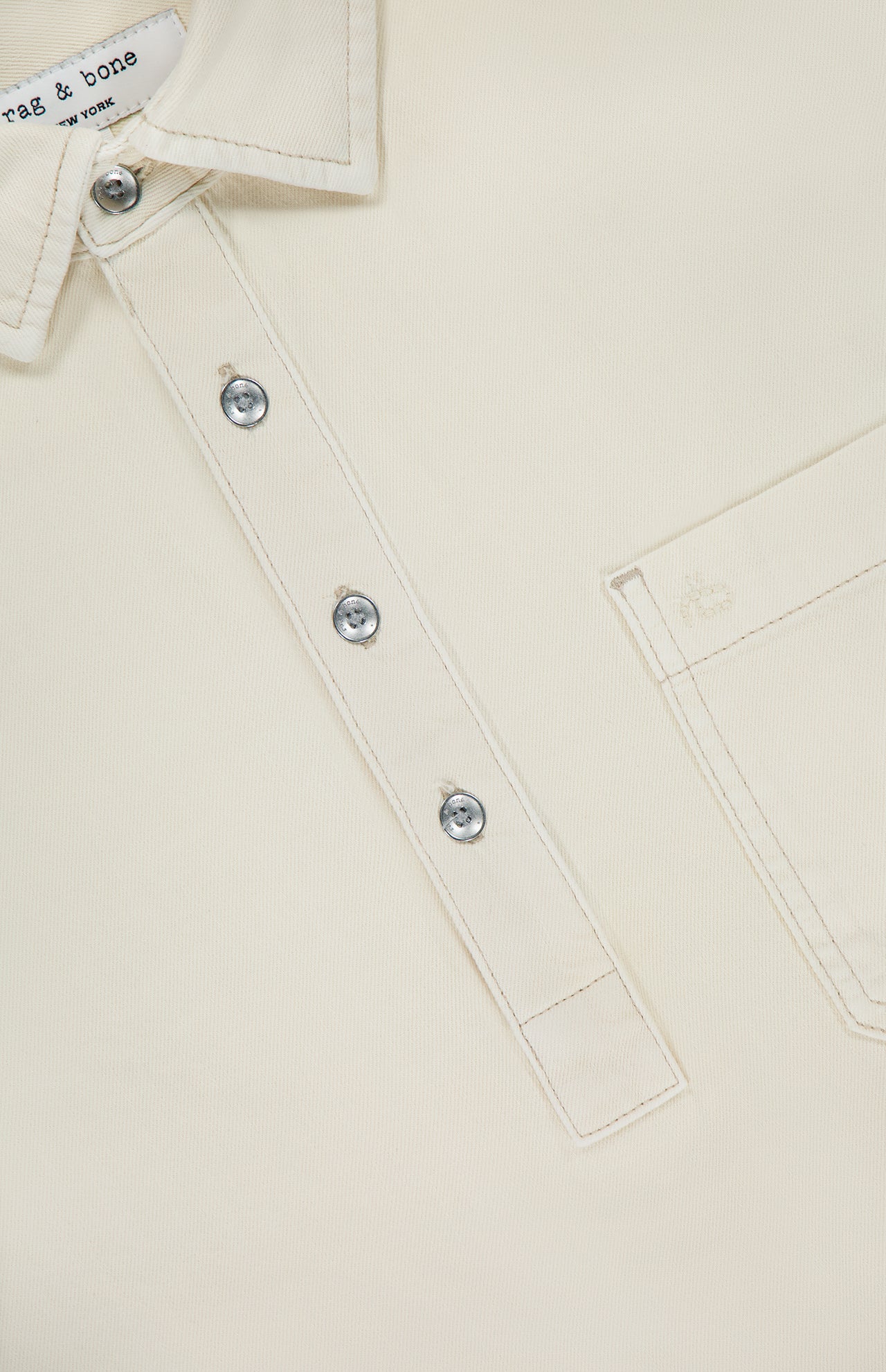 Close-up of RAG & BONE Popover Long Sleeve Shirt in cream cotton, featuring a partial button placket with four silver buttons, right chest pocket, visible white stitching, and a label that reads rag & bone NEW YORK for a minimalist design.