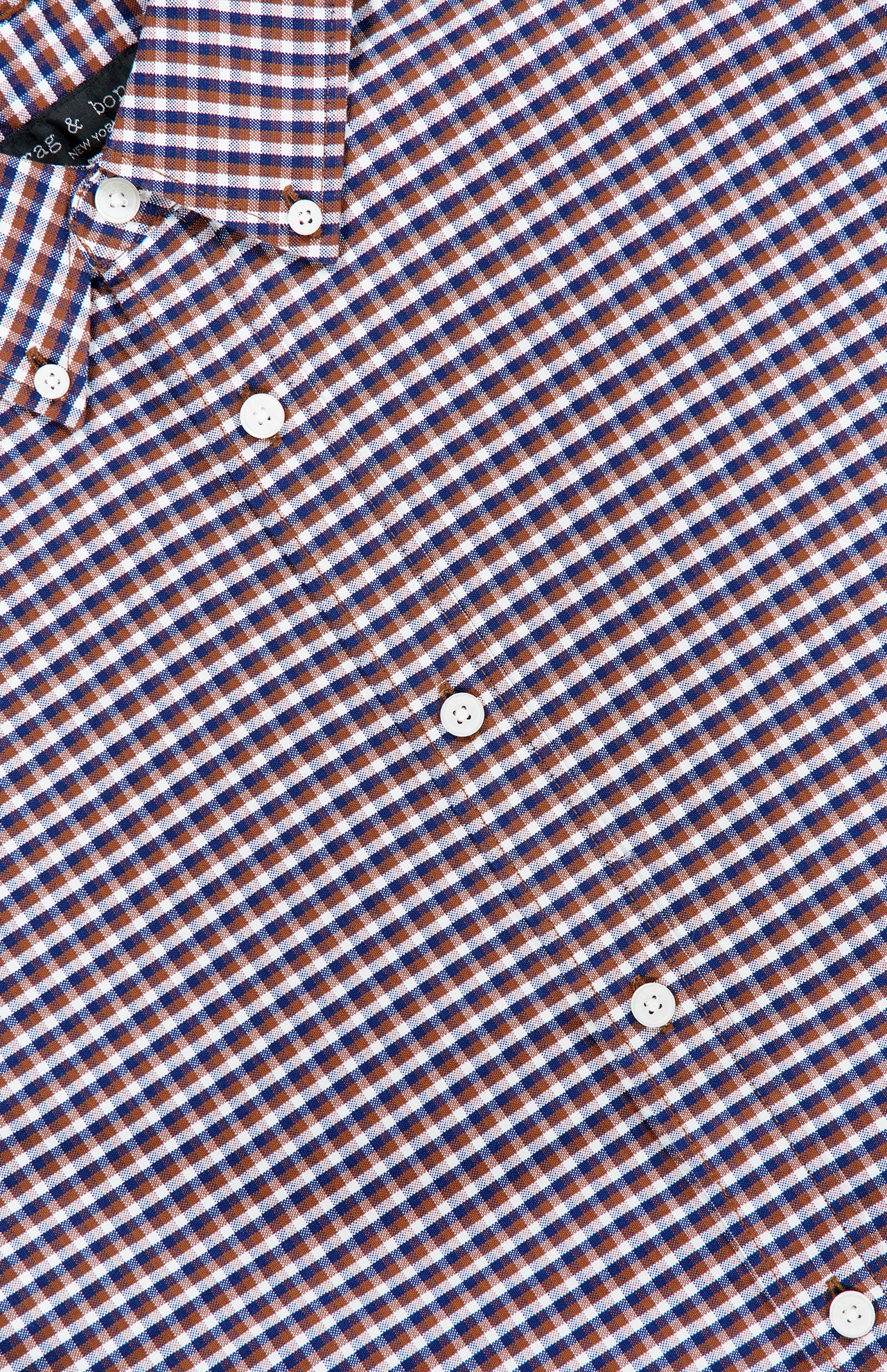 Close-up of the RAG & BONE Classic Oxford Dress Shirt, crafted from 100% cotton with a small brown and navy check pattern accented by white. Crisp white buttons and a structured collar are also visible in the upper left corner.