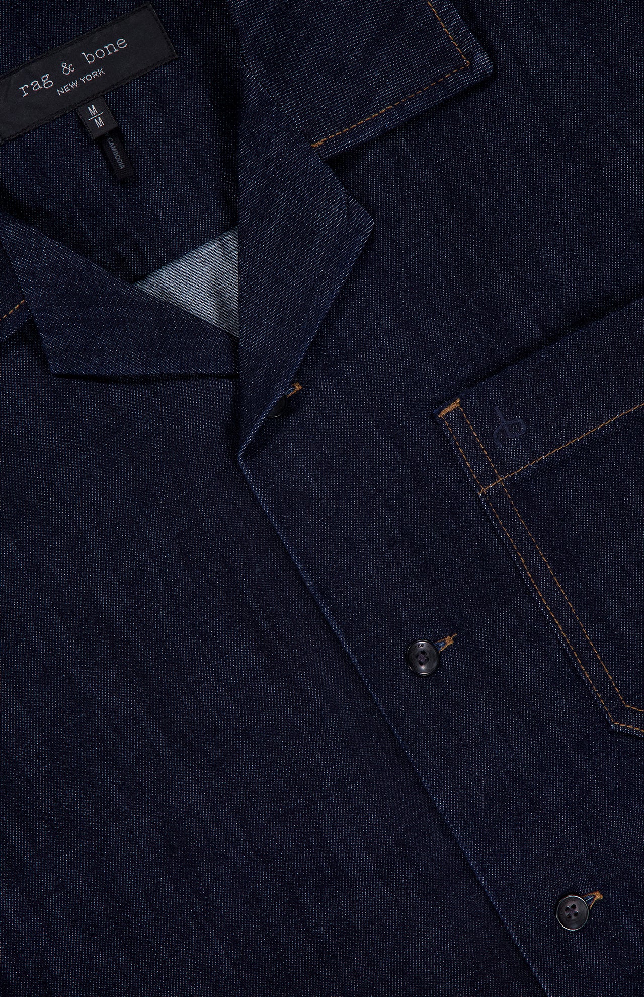 A close-up of the RAG & BONE Resort Denim Shirt features black buttons, orange stitching, a chest pocket, and a branded collar label—an effortless choice for resort style.