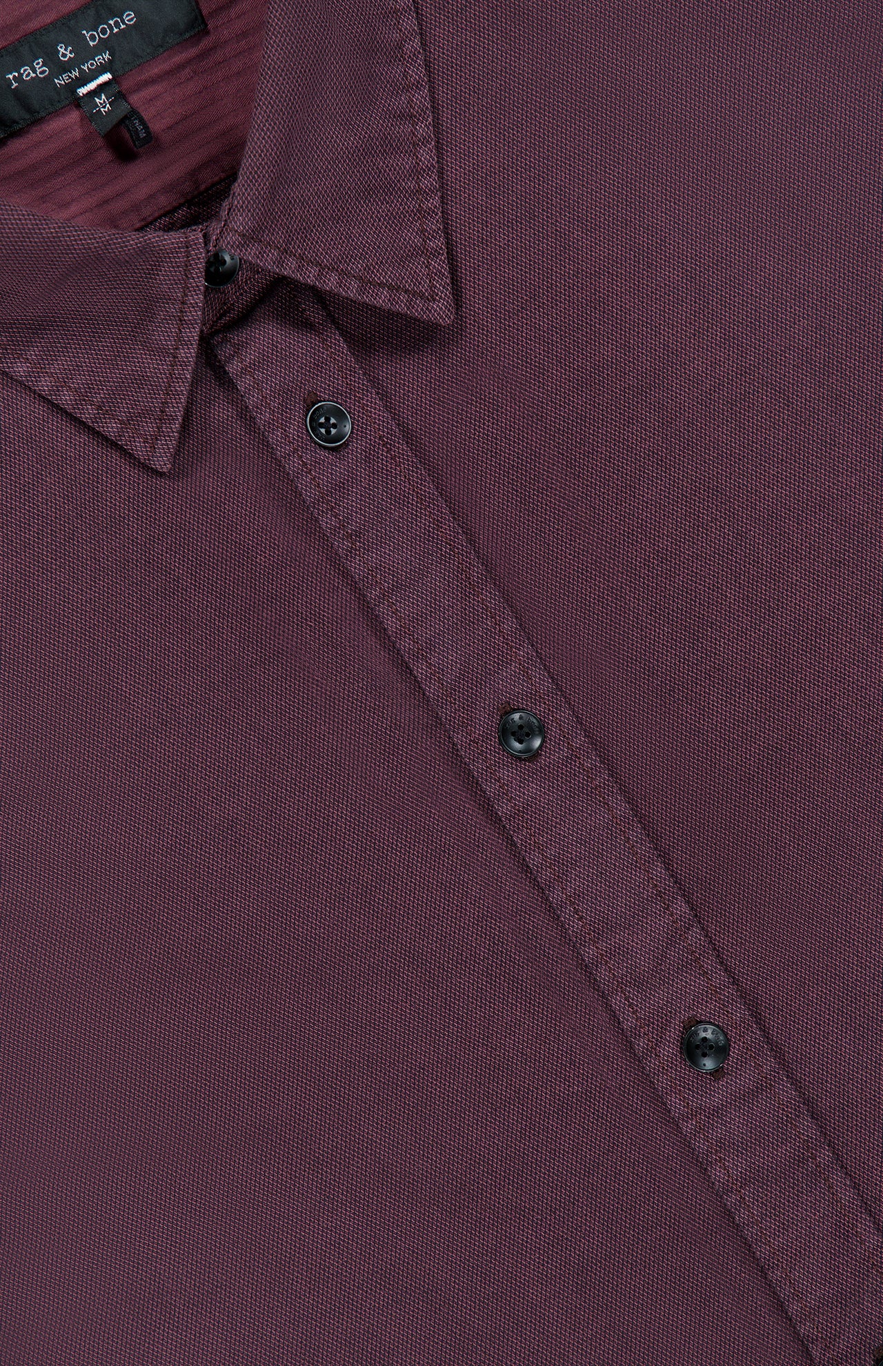 A close-up of the Tomlin Dobby Dress Shirt by RAG & BONE, a burgundy stretch cotton button-up with black buttons and a pointed collar, displayed on a flat surface. The shirt tag displays the brand name RAG & BONE.