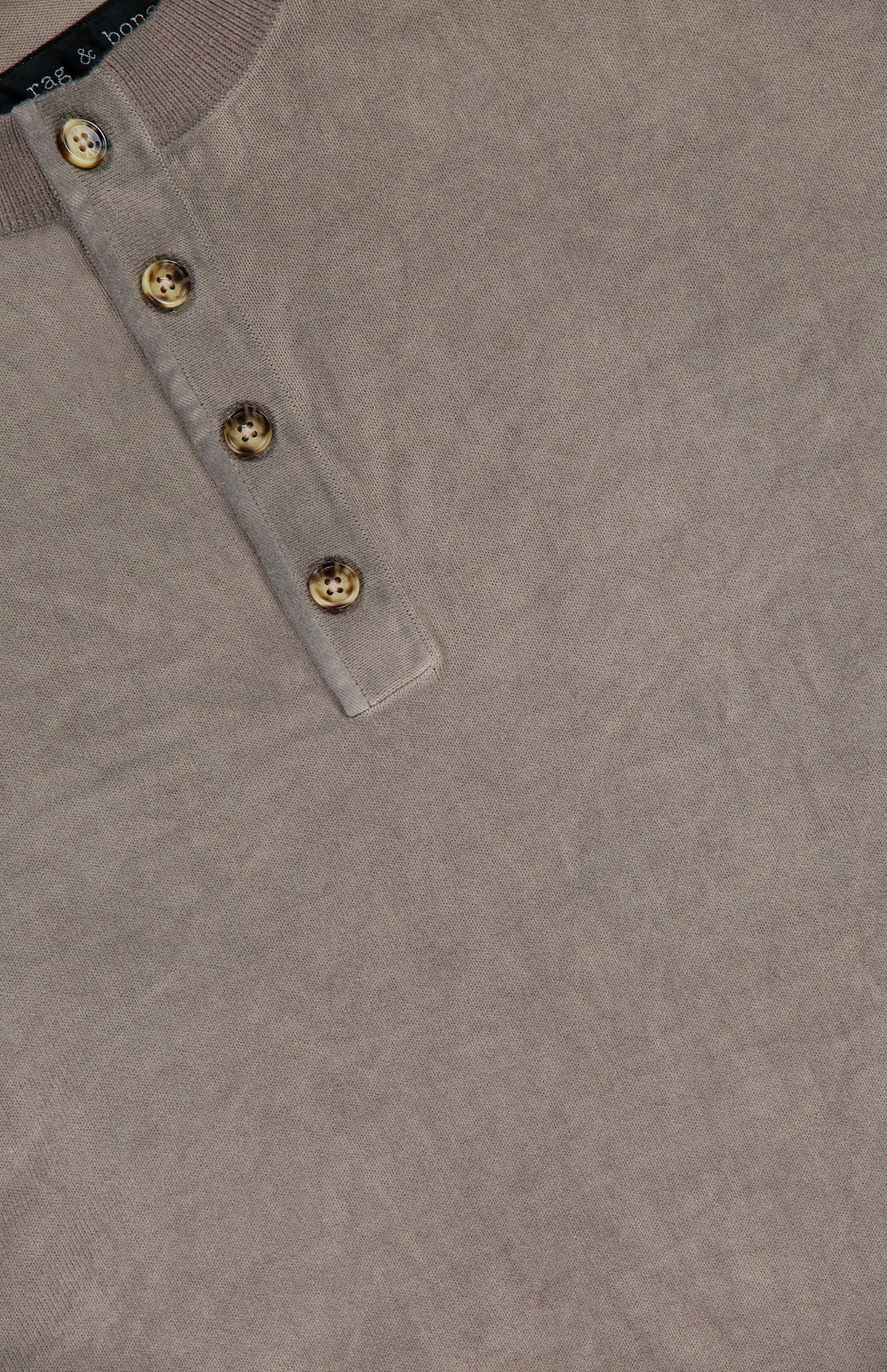 Close-up of the RAG & BONE Washed Cotton Henley Shirt in beige, featuring soft cotton yarn, a partial view of the collar, and four brown buttons along the placket on a textured fabric background.