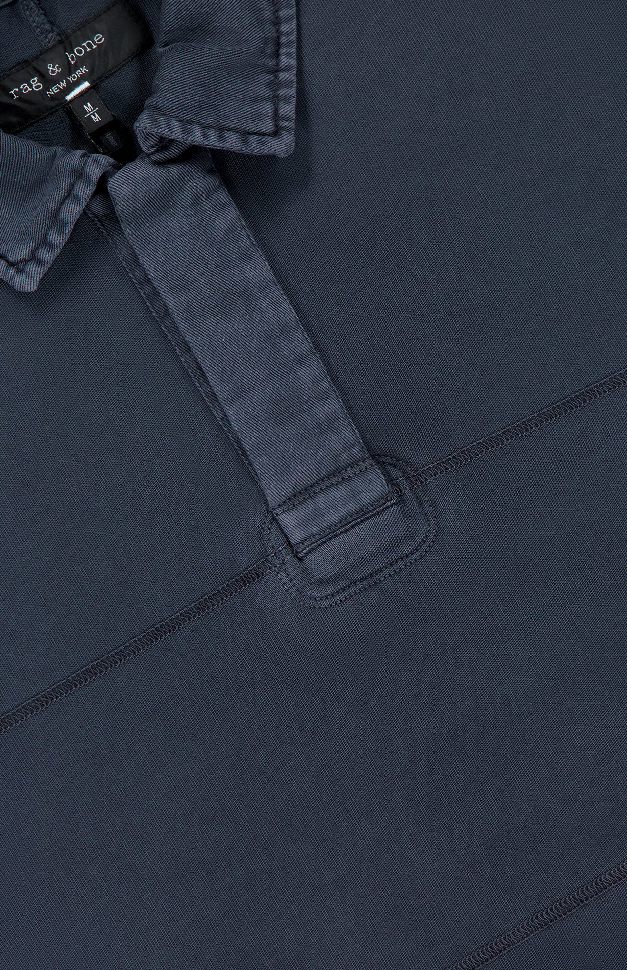 Close-up of the Washed Rugby Long Sleeve Polo Shirt by RAG & BONE in navy blue cotton, featuring a partial placket, visible stitching, and a black "rag & bone new york" collar tag with XL size label.