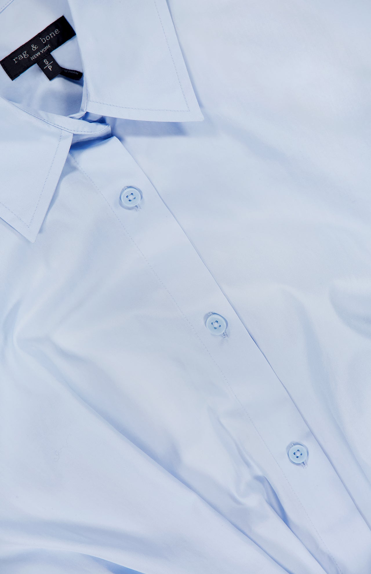 A close-up of the RAG & BONE Andy Pleated Drawcord Top in light blue, featuring a pointed collar and visible buttons, lying flat. The shirt tag displays the brand name.