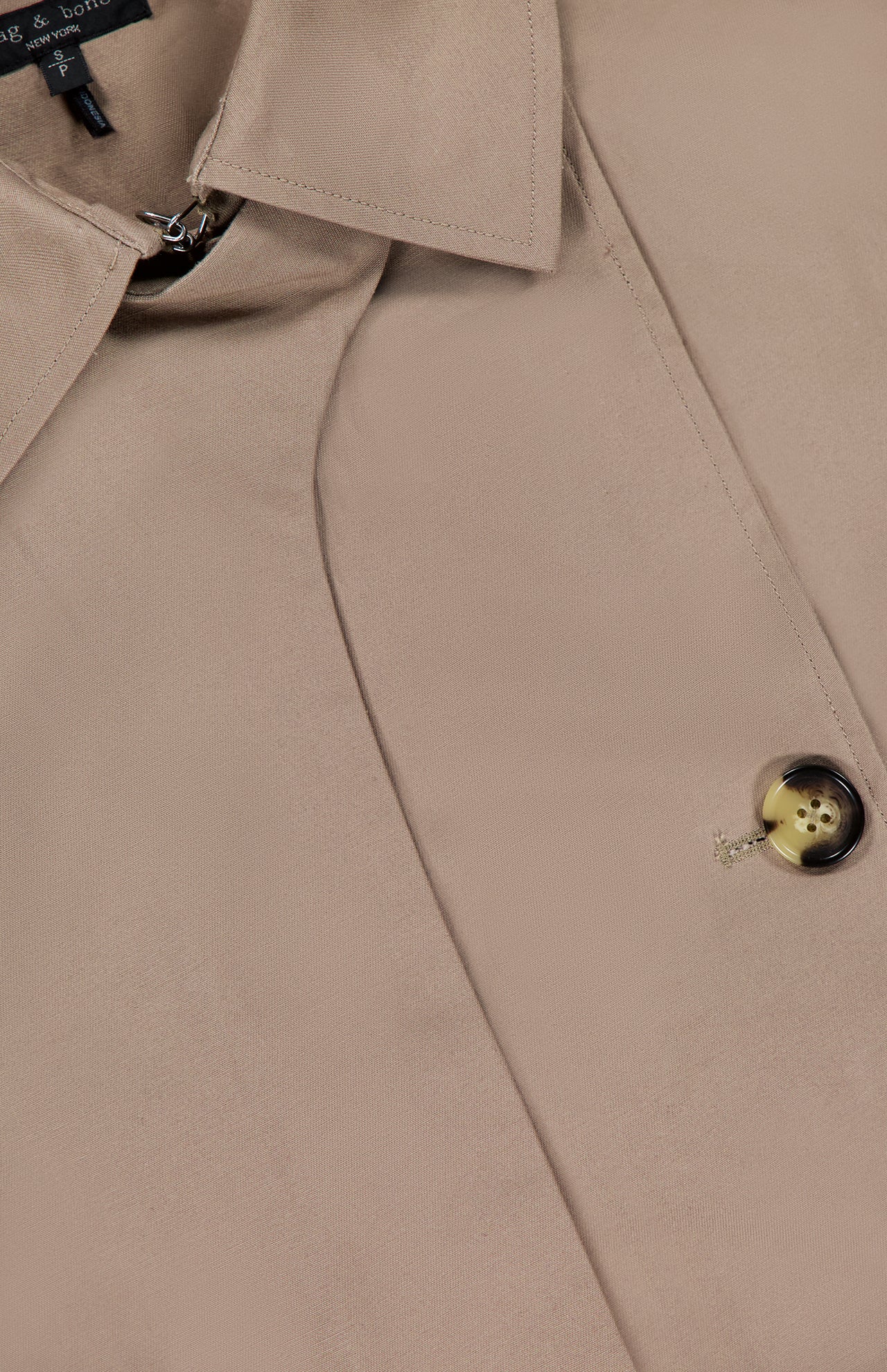 Close-up of the RAG & BONE Jamie Cropped Trench Coat, featuring its collar, partially visible black label, buttonhole, and a single brown button sewn with tan thread—a timeless spring jacket.