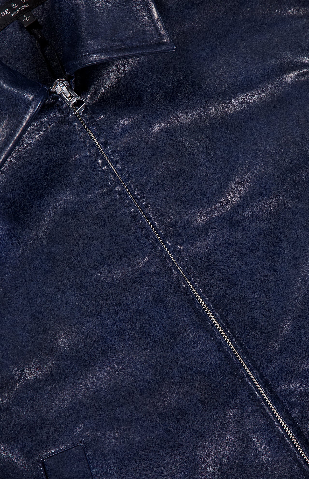 Close-up of the Krista Faux Leather Jacket by RAG & BONE, highlighting the shiny navy blue faux leather texture, a partially unzipped silver zipper, a pointed collar, and part of the front pocket—showcasing the sleek appeal of this modern cropped bomber style.