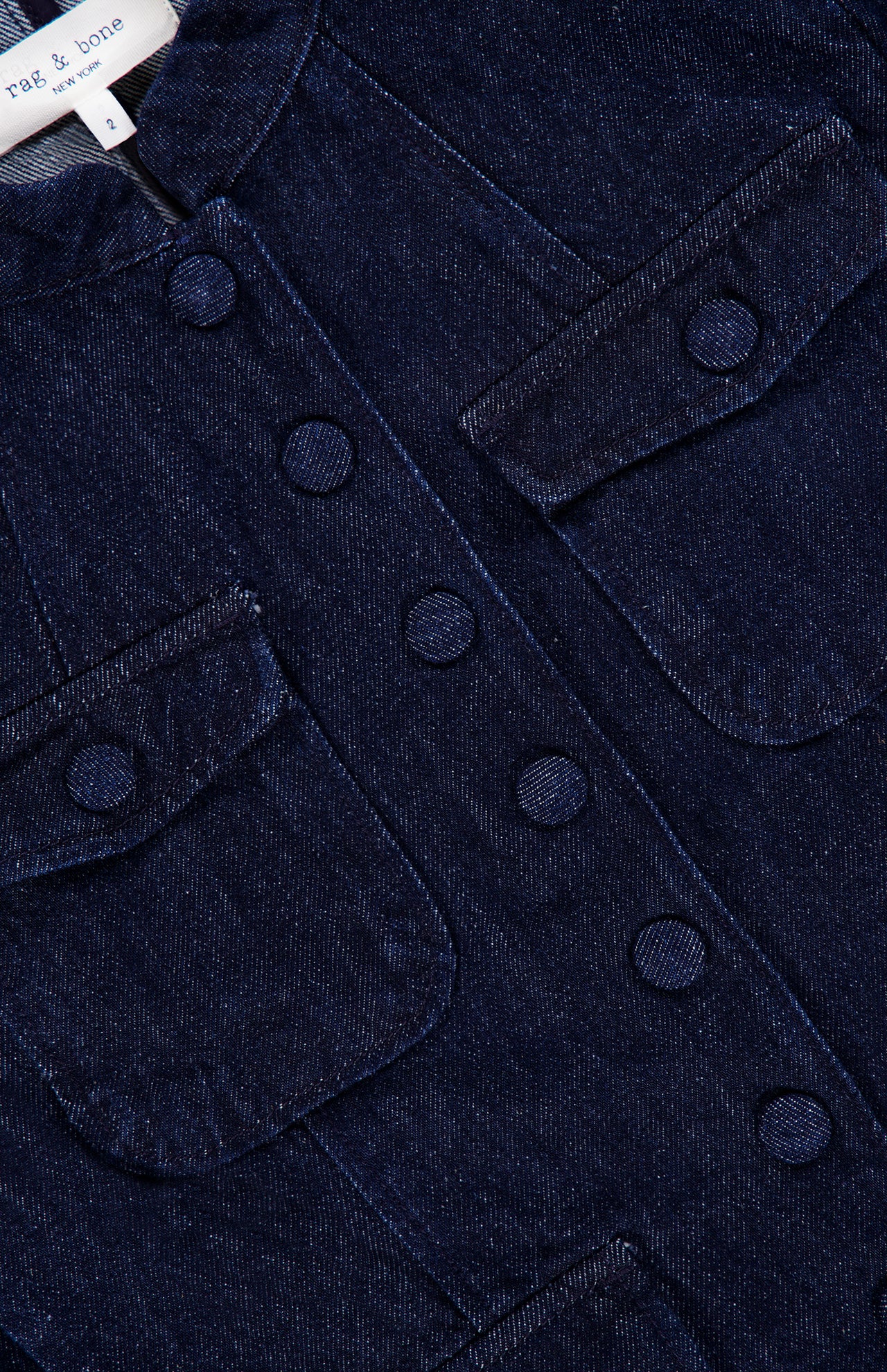 Close-up of the RAG & BONE Mercer Pocketed Denim Blazer in dark blue, showcasing its slim fit design with four flap pockets, round buttons, and visible stitching. A small branded clothing tag is partially visible at the neckline.