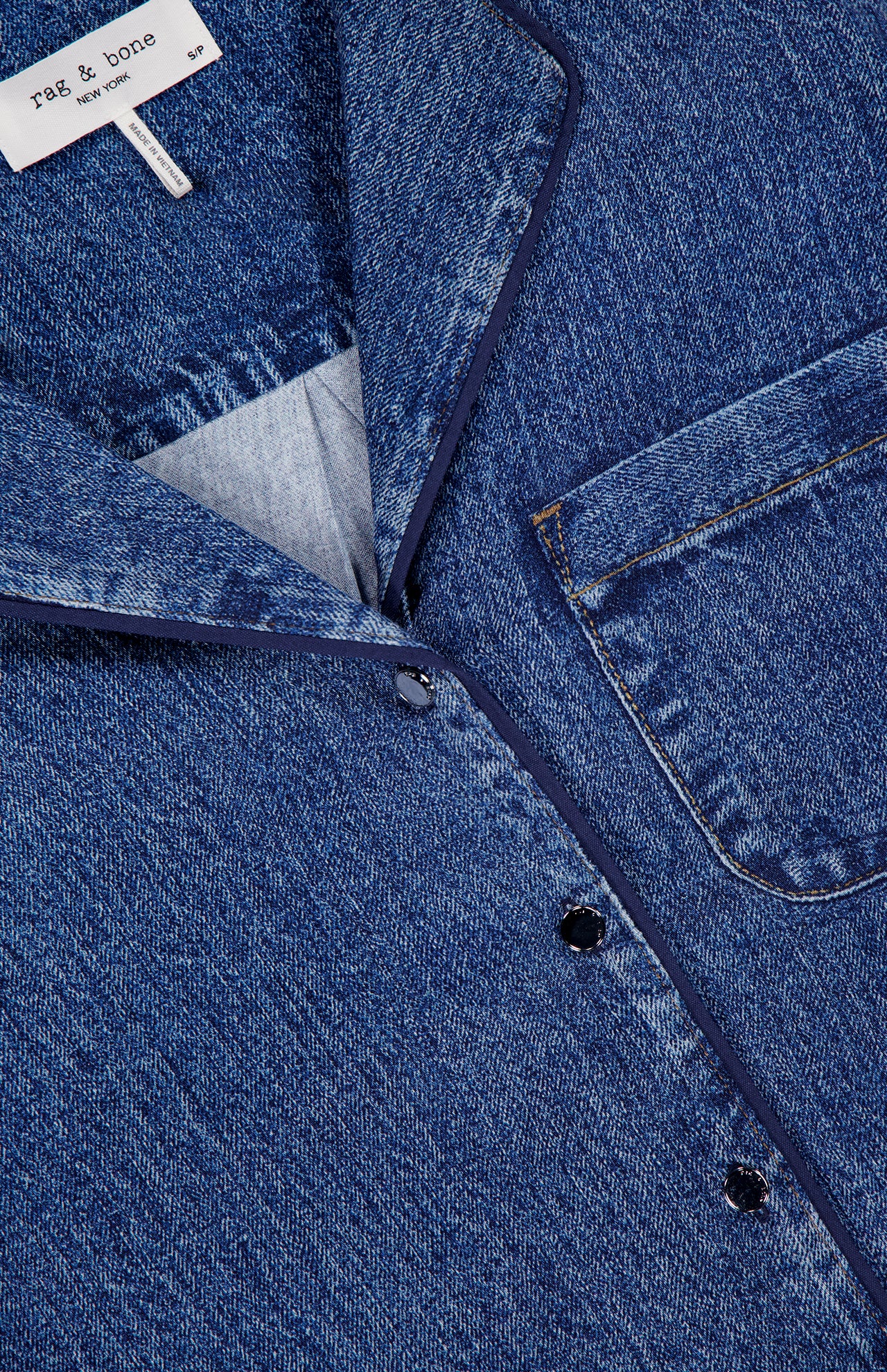 Close-up of the Miramar Slink Short Sleeve Lounge Shirt by RAG & BONE in blue denim-look drapey fabric, featuring a chest pocket, classic collar, contrast stitching, dark buttons, and a visible brand label on the collar.