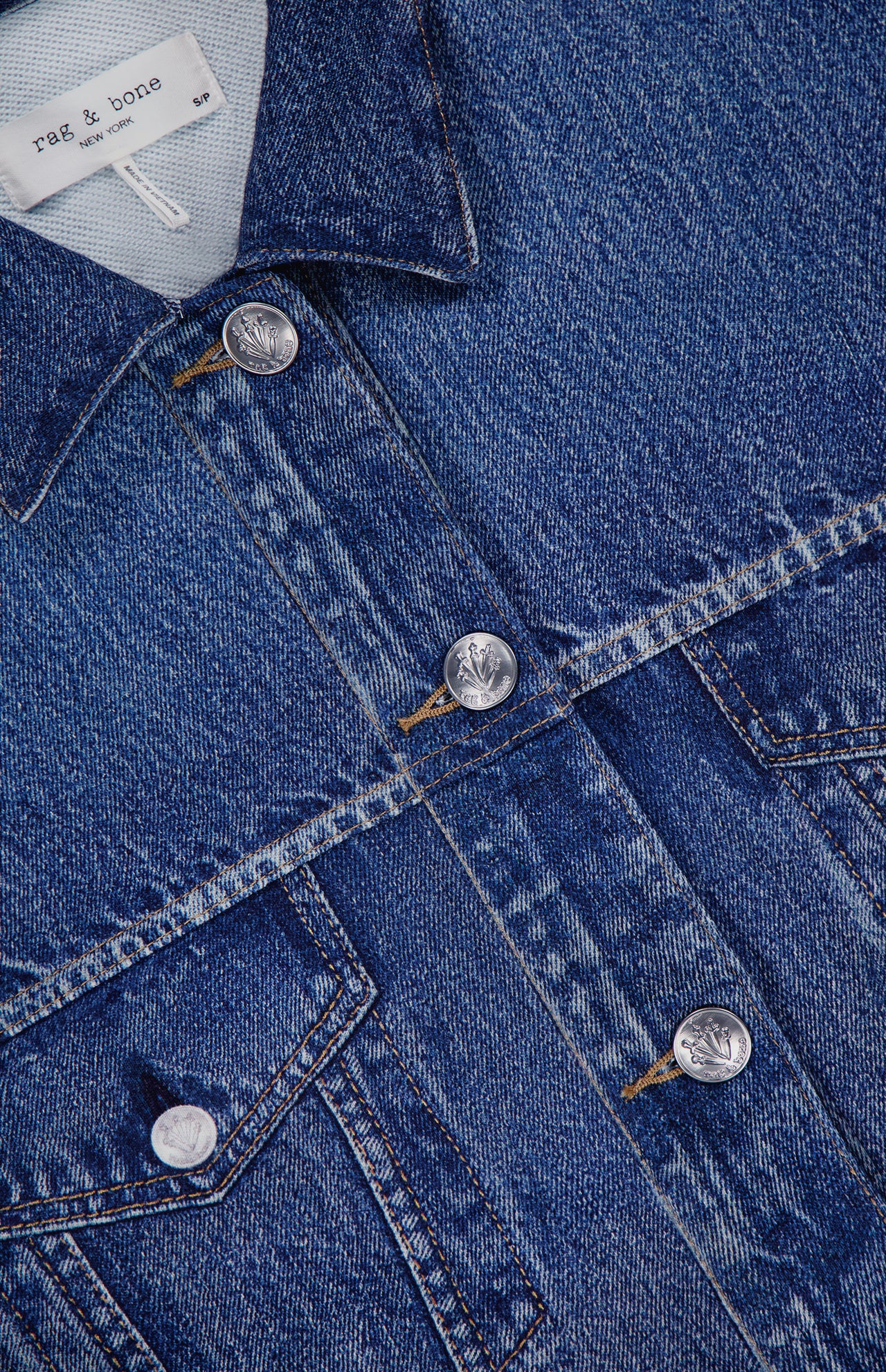 Close-up of the Miramar Terry Trucker Jacket by RAG & BONE, a blue denim-look jacket with flap pockets, silver buttons, visible stitching, and a rag & bone label inside the collar.
