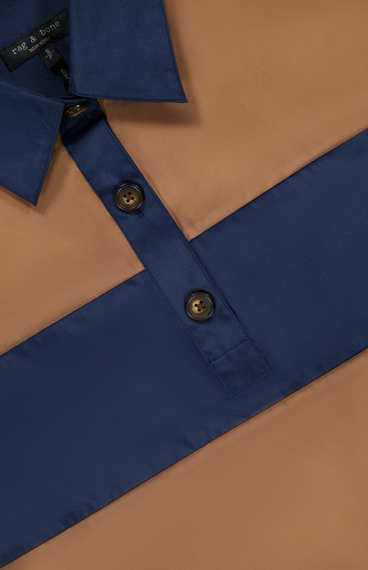 A close-up of the RAG & BONE Oversized Tie Hem Rugby Shirt, showcasing its tan and navy thick horizontal stripes, contrasting blue collar and button placket with two visible brown buttons, and classic long-sleeved polo styling.