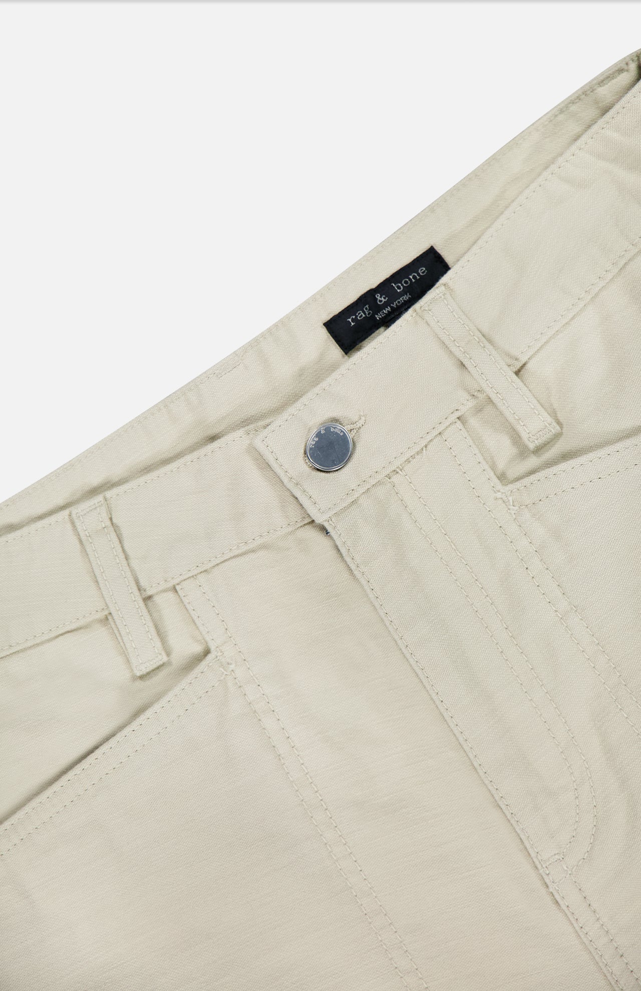 Close-up of the waist area of Stevie Utility Tapered Pant in light tan, 100% cotton, showing front button, belt loops, and front pockets. Inside the waistband is a black RAG & BONE label.