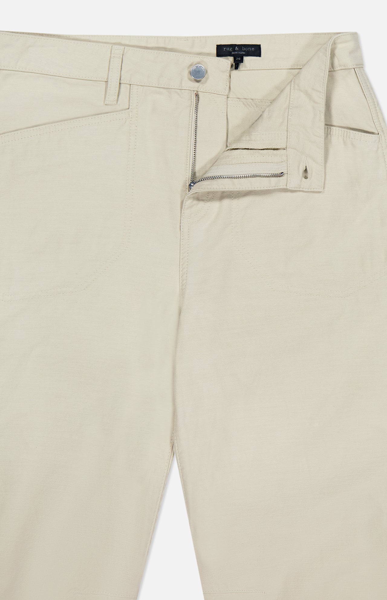 The Stevie Utility Tapered Pant by RAG & BONE is a light tan, utility-inspired pair featuring a visible zipper fly, button closure, belt loops, front pockets, and a partially open waistband showing the inside label.