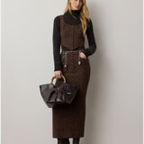 A woman stands against a plain wall, wearing the RALPH LAUREN COLLECTION Cashmere Turtleneck Long Sleeve Sweater in black, paired with a brown vest, a matching long skirt, and heeled boots. She carries a dark leather handbag and her long hair is worn loose.