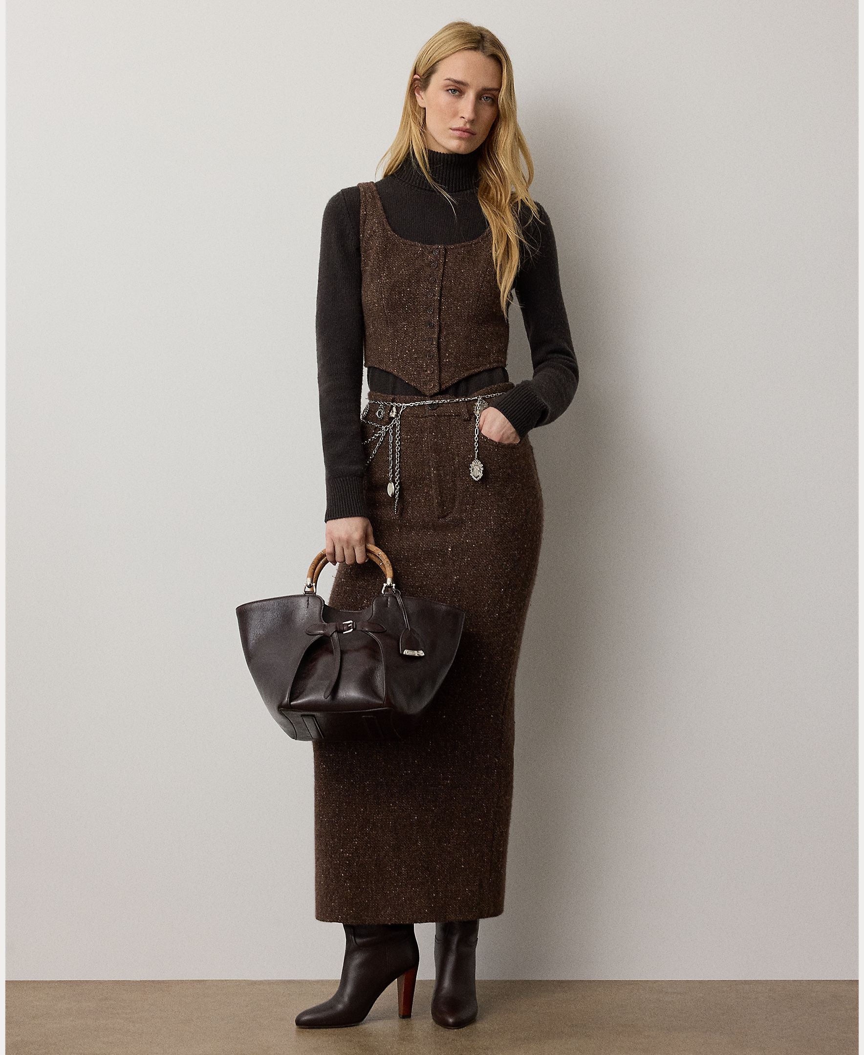 A woman stands against a plain wall, wearing the RALPH LAUREN COLLECTION Cashmere Turtleneck Long Sleeve Sweater in black, paired with a brown vest, a matching long skirt, and heeled boots. She carries a dark leather handbag and her long hair is worn loose.