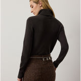 A woman with blonde hair, seen from behind against a plain light background, wears a brown skirt and the RALPH LAUREN COLLECTION Cashmere Turtleneck Long Sleeve Sweater. A decorative chain belt accents her waist, while the luxurious Italian-made sweater adds an elegant touch to her refined style.
