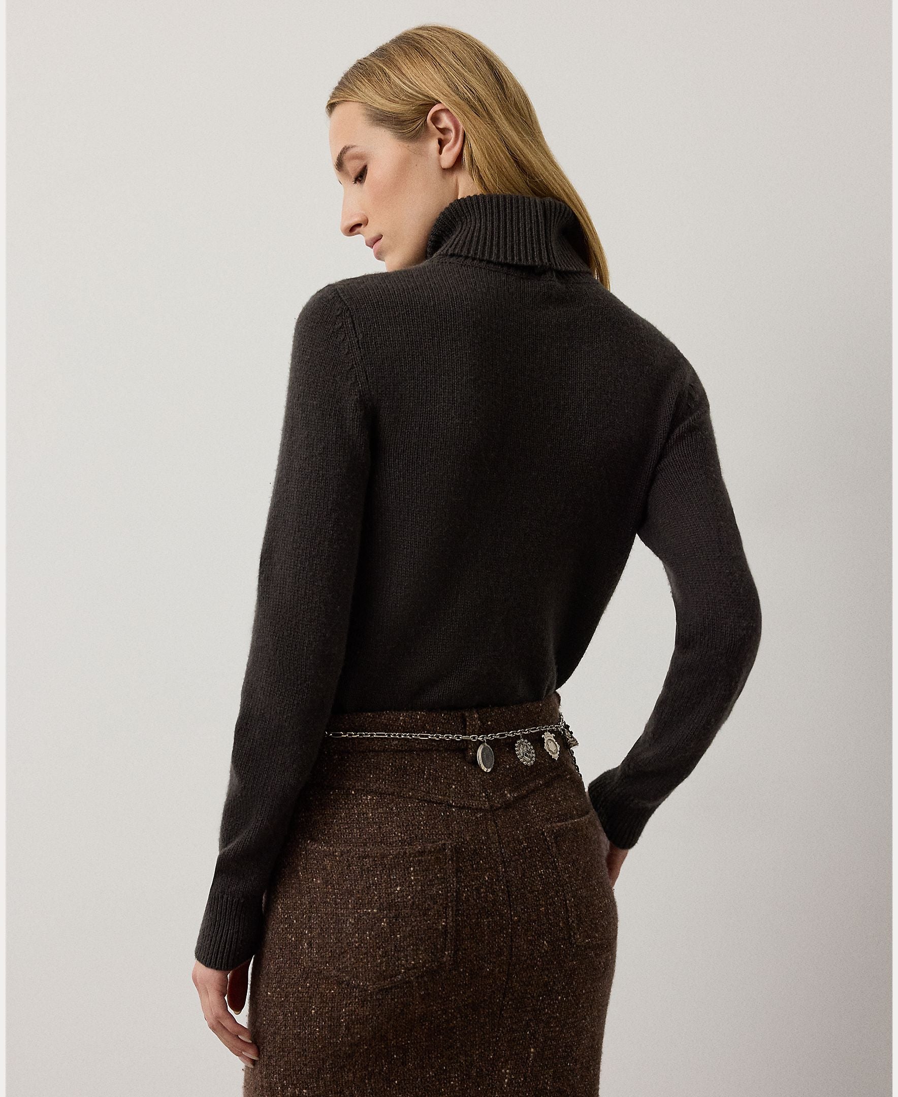 A woman with blonde hair, seen from behind against a plain light background, wears a brown skirt and the RALPH LAUREN COLLECTION Cashmere Turtleneck Long Sleeve Sweater. A decorative chain belt accents her waist, while the luxurious Italian-made sweater adds an elegant touch to her refined style.