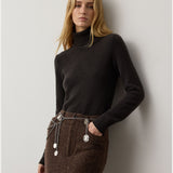 A woman with long blonde hair wears the RALPH LAUREN COLLECTION Cashmere Turtleneck Long Sleeve Sweater, paired with a brown skirt and a silver chain belt with medallions, standing against a plain light gray wall.