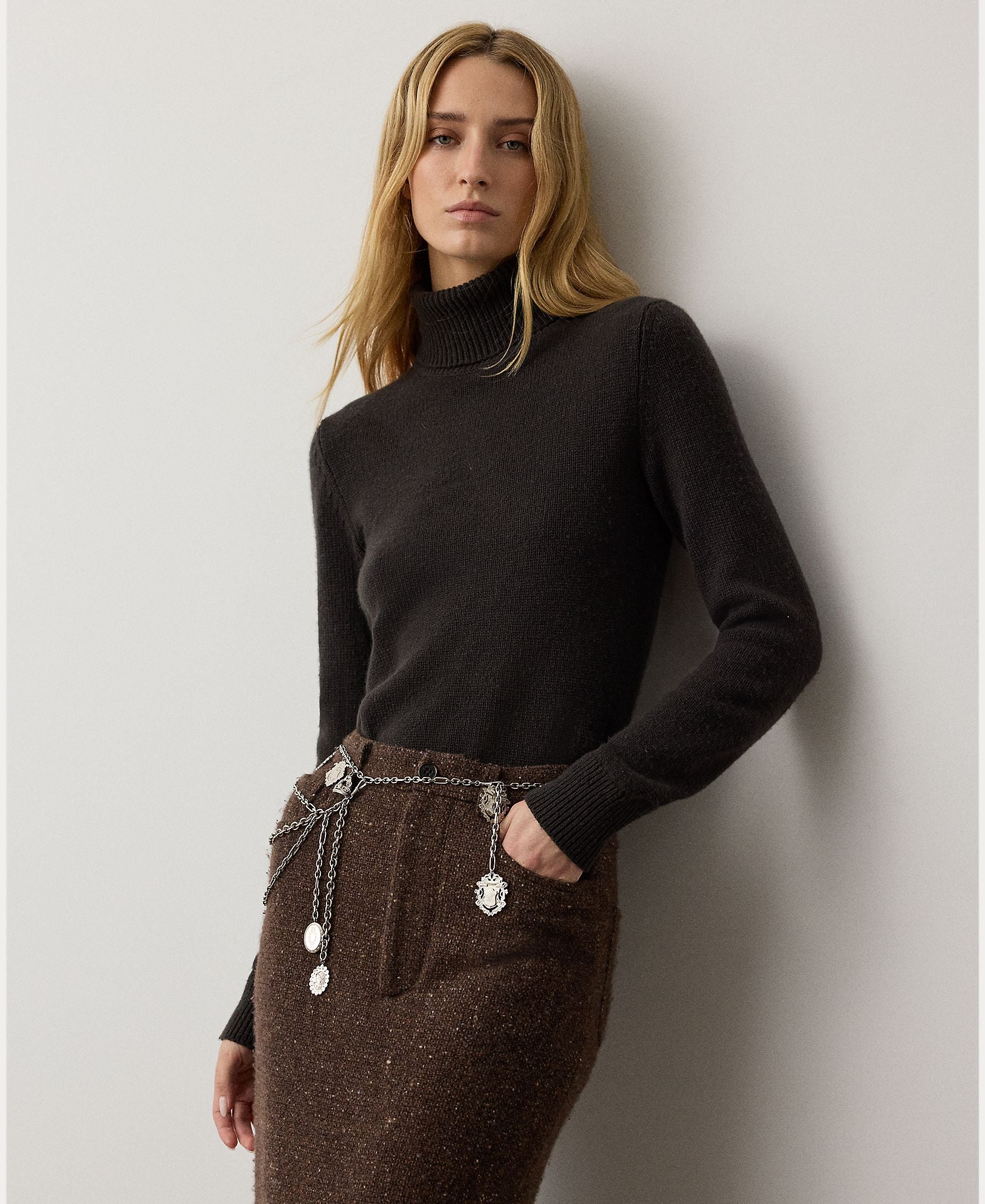 A woman with long blonde hair wears the RALPH LAUREN COLLECTION Cashmere Turtleneck Long Sleeve Sweater, paired with a brown skirt and a silver chain belt with medallions, standing against a plain light gray wall.