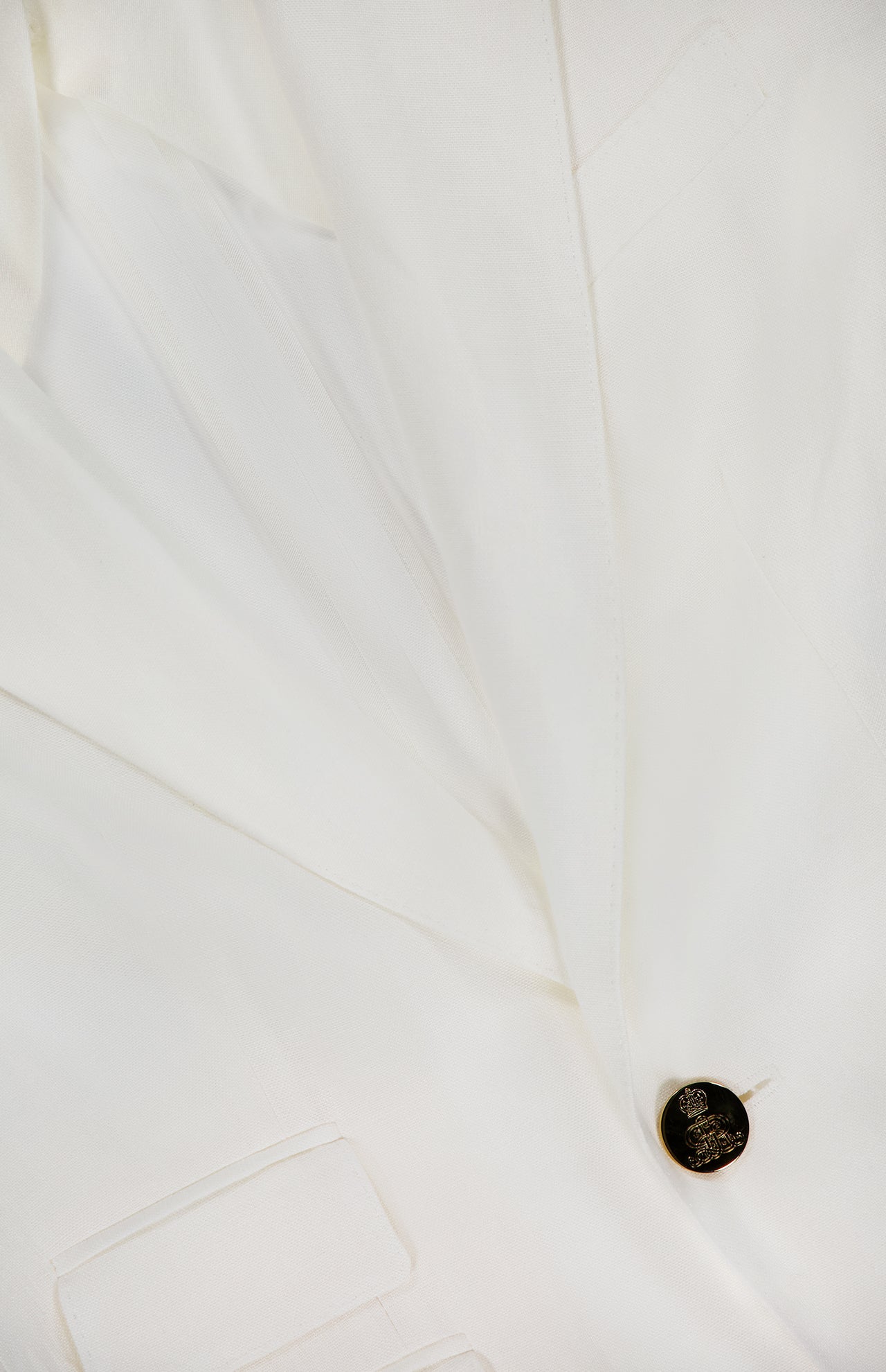 Close-up of the Classic Linen Parker Blazer by RALPH LAUREN COLLECTION, an off-white piece with two pockets, a lapel, and a single dark crest-embossed button. The linen fabric looks smooth and crisp.