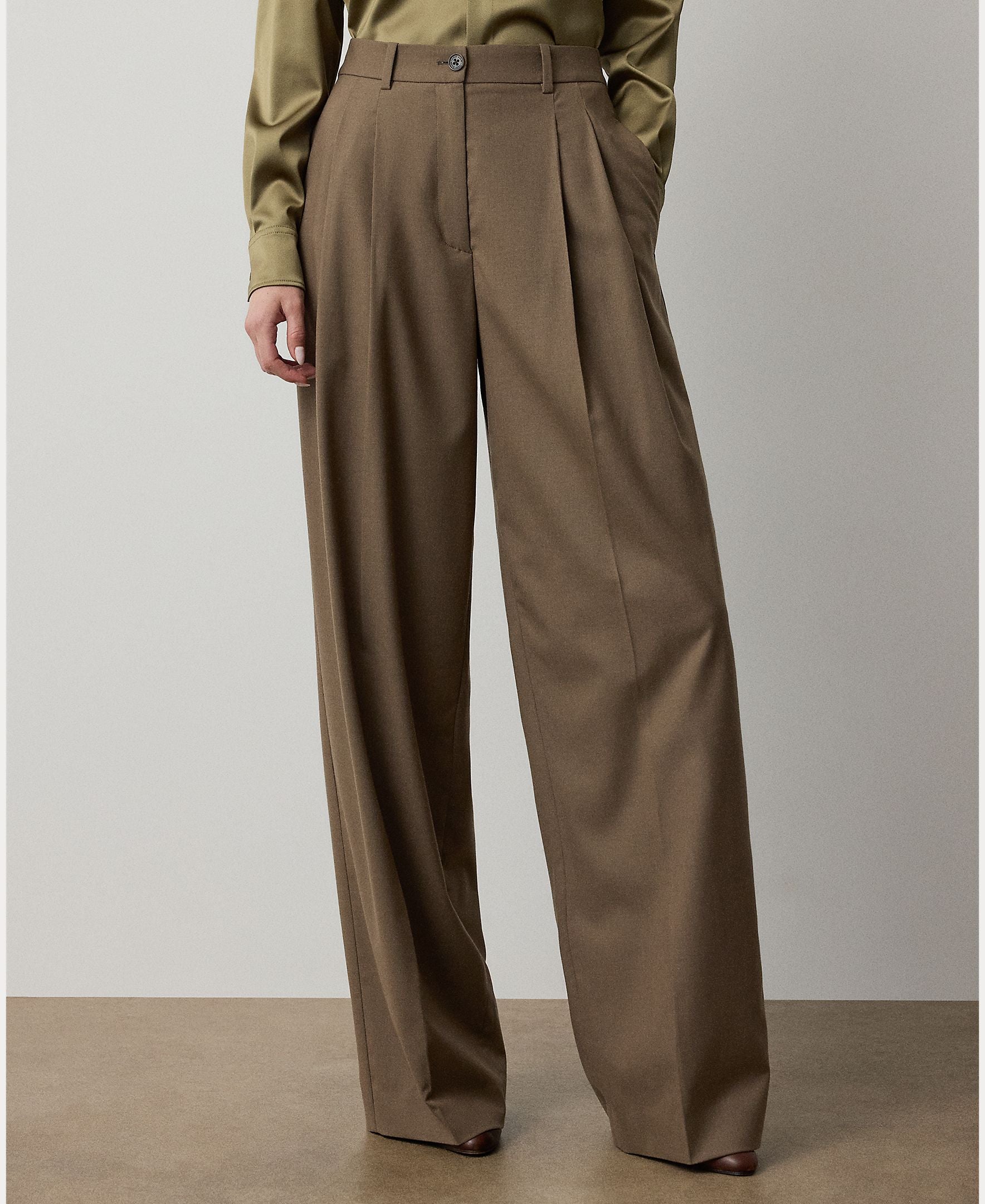 A person is wearing the Fine Wool Standard Pants from RALPH LAUREN COLLECTION—relaxed fit, wide-leg, high-waisted brown trousers in stretch-wool with pleats—paired with an olive green long-sleeve shirt. The photo is cropped below the shoulders, focusing on the lower body.
