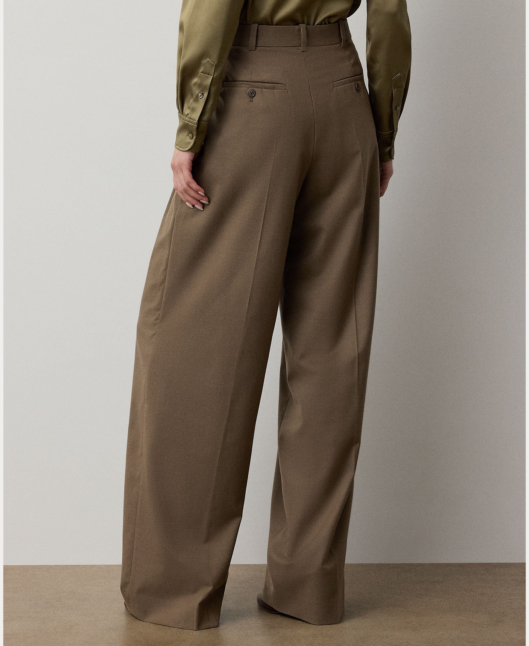 A person wearing the RALPH LAUREN COLLECTION Fine Wool Standard Pants in a relaxed, wide-leg brown style and an olive-green blouse stands with their back to the camera against a neutral background.
