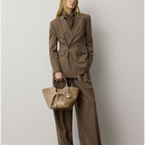 A woman stands against a plain wall, exuding modern sophistication in a relaxed fit as she dons the Fine Wool Standard Pants from the RALPH LAUREN COLLECTION—a pair of wide-leg brown trousers crafted from stretch-wool fabric and styled with a double-breasted suit. She accessorizes with a textured brown handbag and gazes thoughtfully to the side.