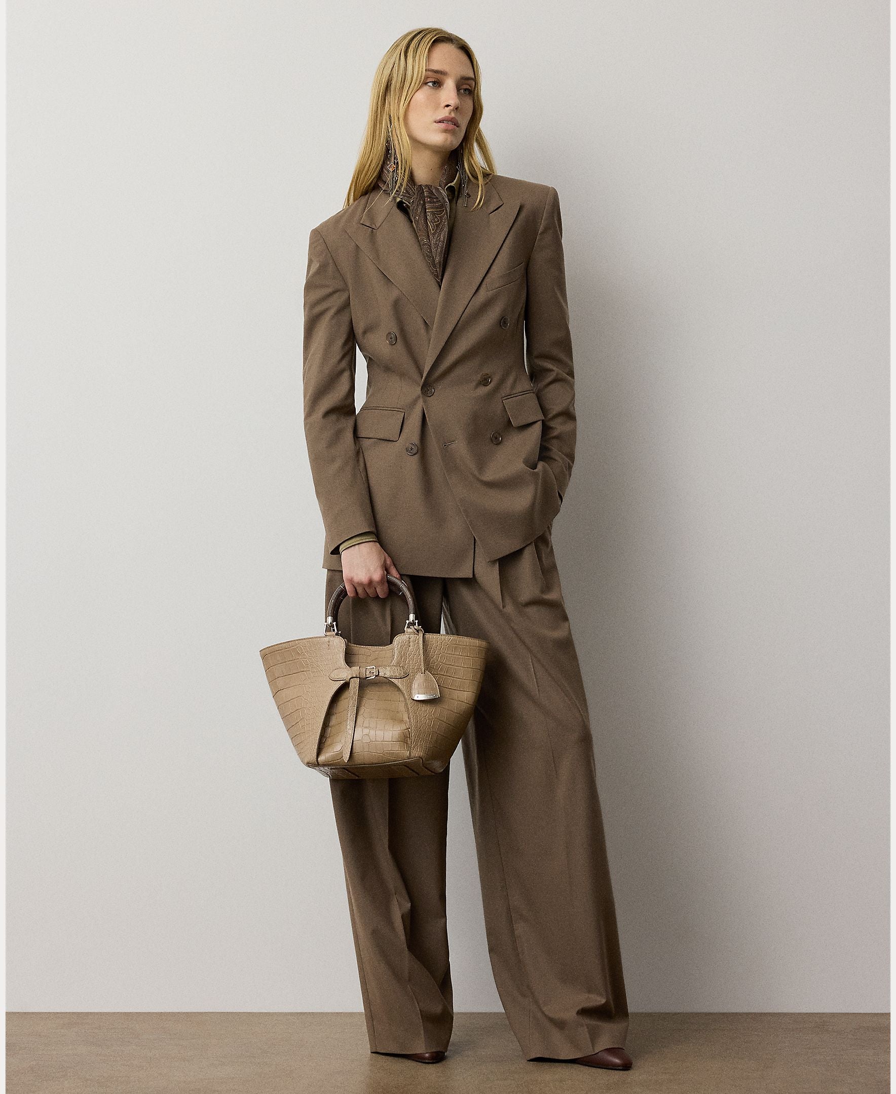 A woman stands against a plain wall, exuding modern sophistication in a relaxed fit as she dons the Fine Wool Standard Pants from the RALPH LAUREN COLLECTION—a pair of wide-leg brown trousers crafted from stretch-wool fabric and styled with a double-breasted suit. She accessorizes with a textured brown handbag and gazes thoughtfully to the side.