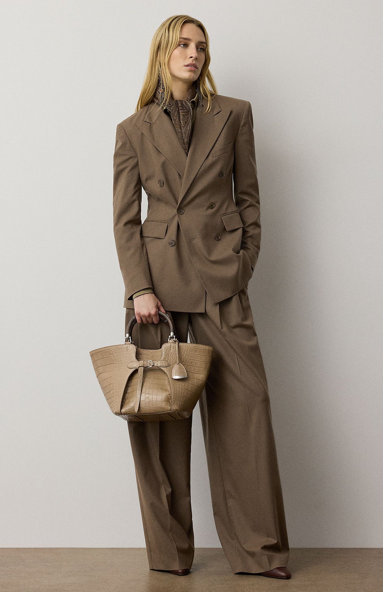 A woman in a brown double-breasted blazer and relaxed fit RALPH LAUREN COLLECTION Fine Wool Standard Pants holds a textured beige handbag.