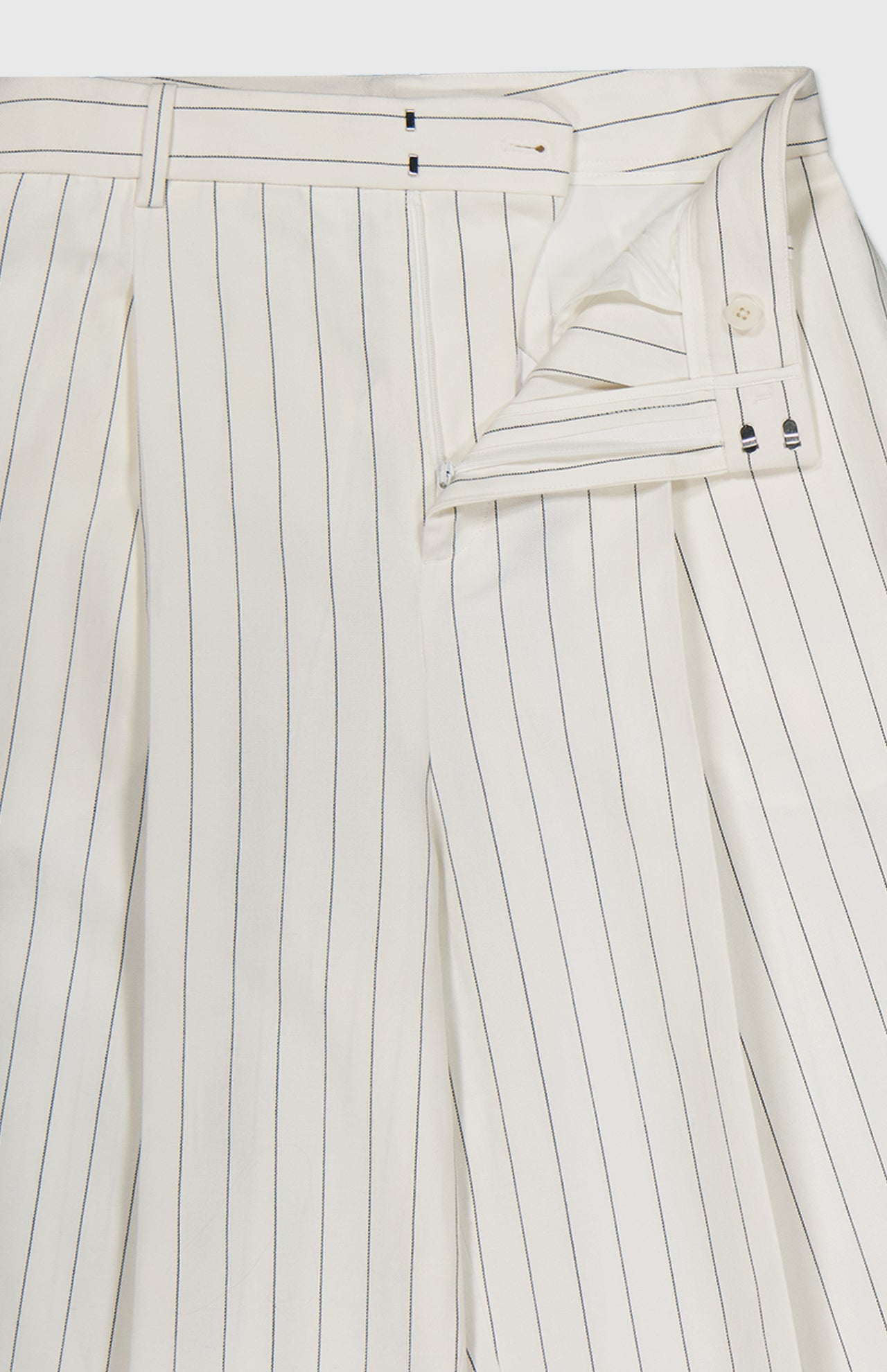 The Harlo Pleated Stripe Suit Pants by RALPH LAUREN COLLECTION feature a carrot silhouette in off-white with thin black pinstripes, crafted from pleated cotton-blend suiting and shown partially unbuttoned to reveal detailed inner buttons.
