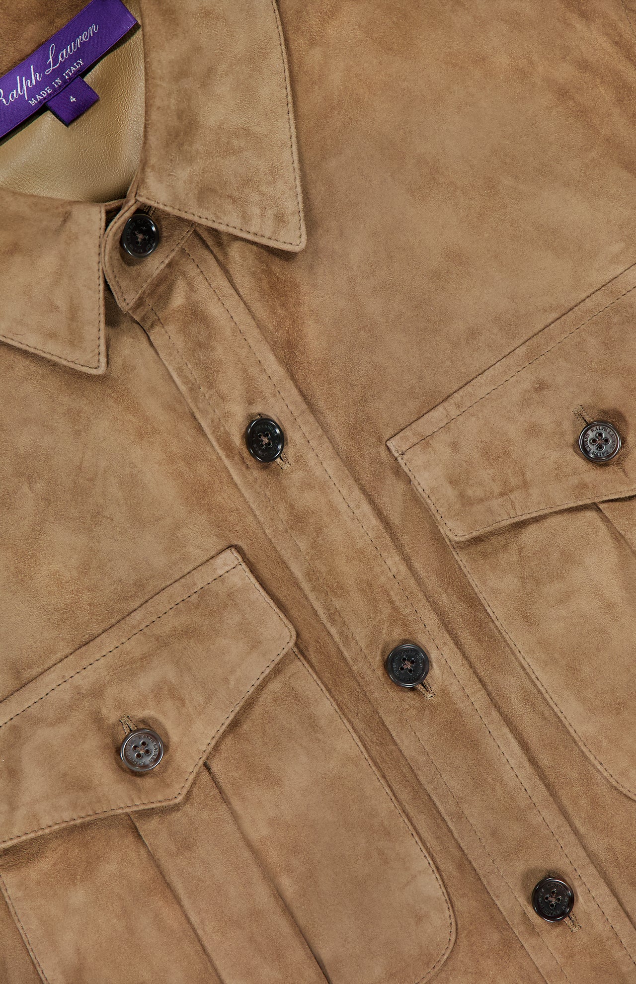 A close-up of the Landry Lightweight Suede Long Sleeve Shirt from the RALPH LAUREN COLLECTION showcases its light brown suede fabric, black buttons, and flap chest pockets. The visible collar tag features "Ralph Lauren" in white script on a purple background, emphasizing the refined details of this luxury shirt.