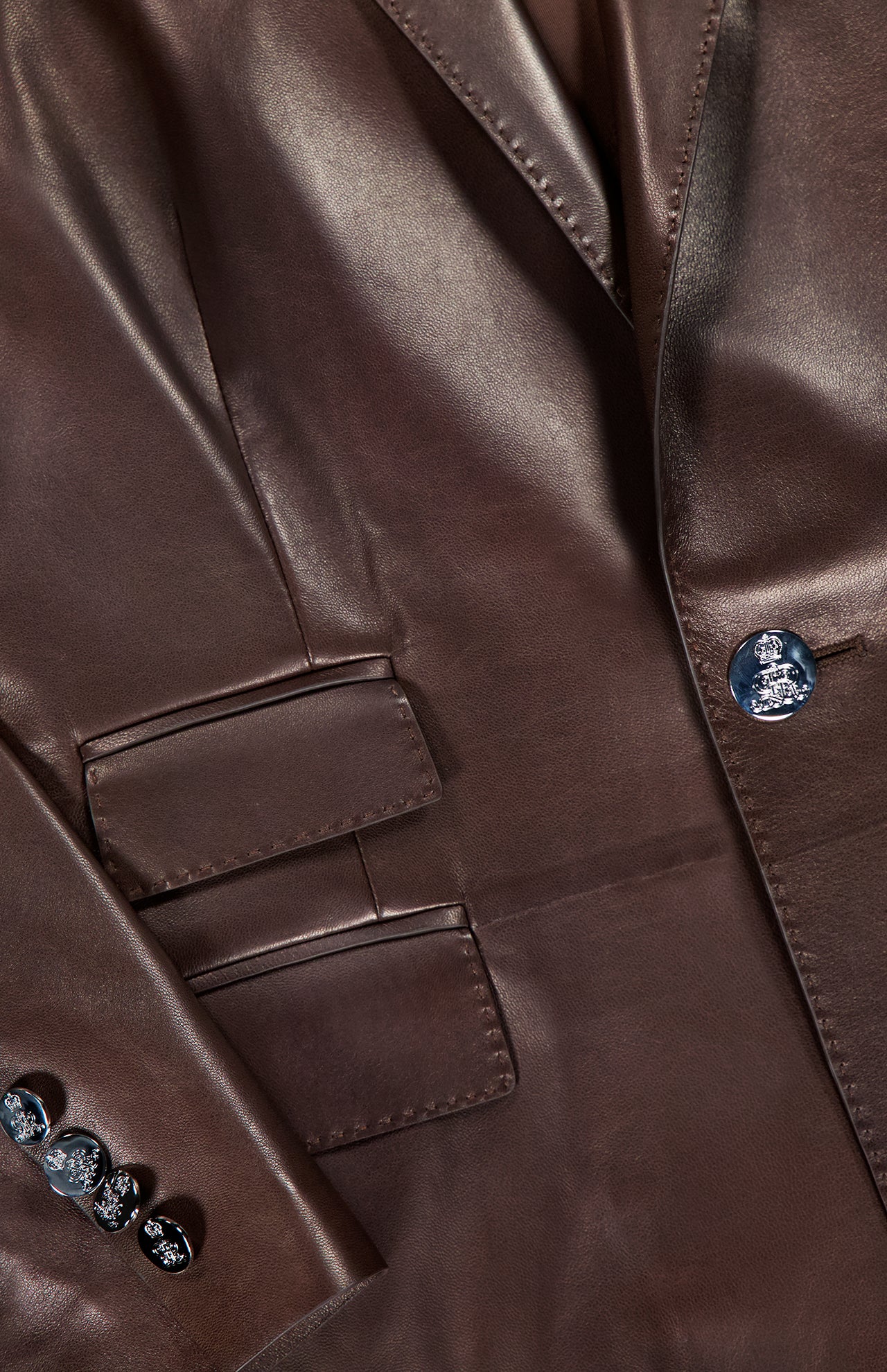 Close-up of the Parker Leather Slim Fit Blazer by RALPH LAUREN COLLECTION, featuring chocolate brown smooth, glossy leather, sharp stitching, two flap pockets, and dark buttons on the sleeve and front.