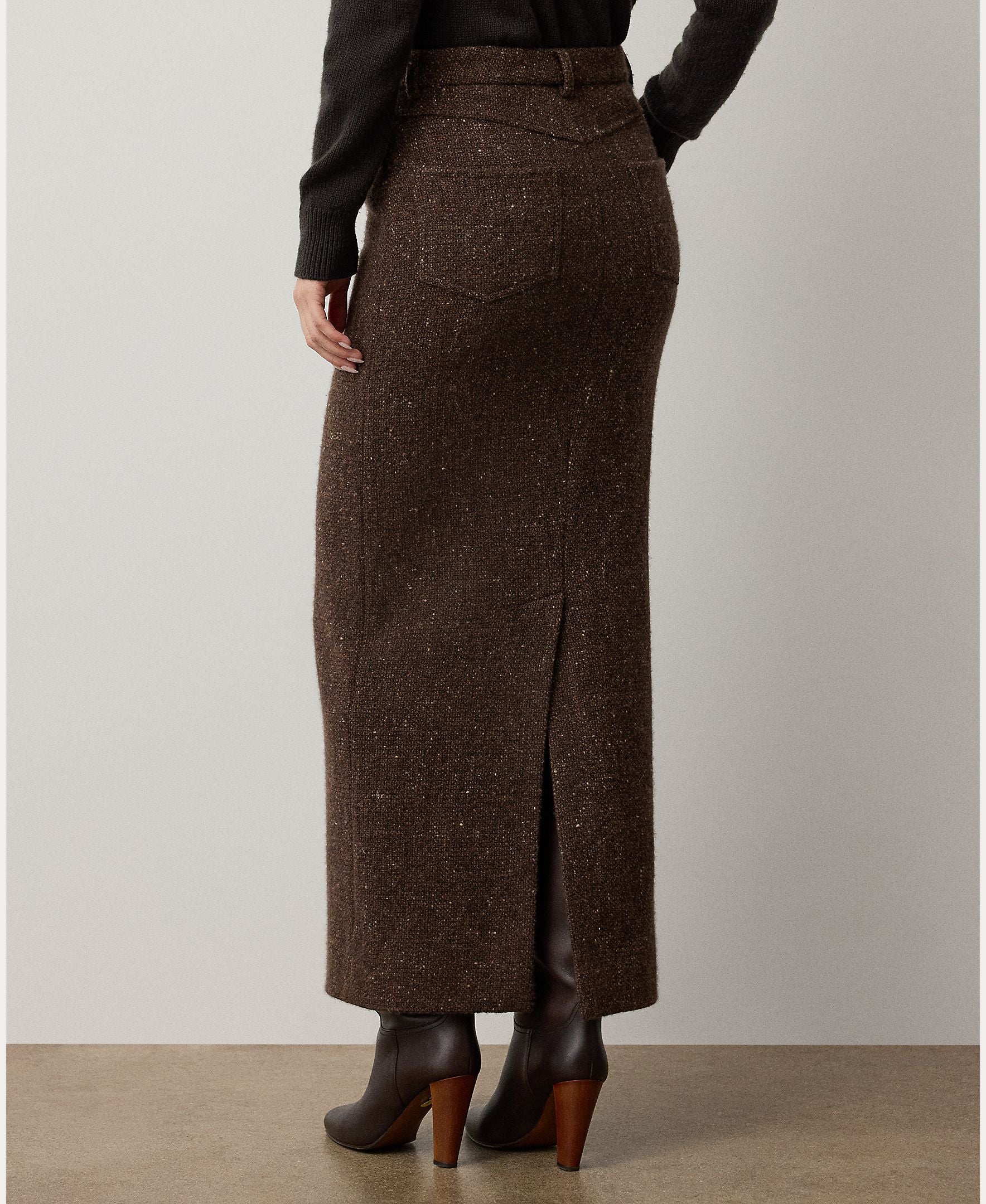 A person stands facing away from the camera against a plain background, wearing the RALPH LAUREN COLLECTION Tweed Ankle Pencil Skirt in long brown wool-cashmere with a back slit, paired with a black top and brown heeled boots.