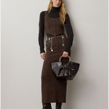 A woman stands against a plain wall, wearing the RALPH LAUREN COLLECTION Tweed Ankle Pencil Skirt paired with a matching vest, a black turtleneck, heeled boots, a metallic belt, and holding a dark handbag with round handles.