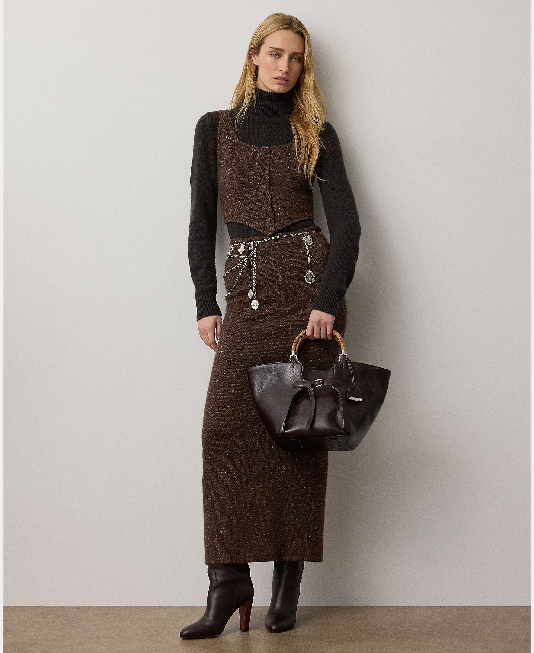 A woman stands against a plain wall, wearing the RALPH LAUREN COLLECTION Tweed Ankle Pencil Skirt paired with a matching vest, a black turtleneck, heeled boots, a metallic belt, and holding a dark handbag with round handles.