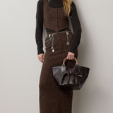 A woman wears a brown tweed vest and a matching RALPH LAUREN COLLECTION Tweed Ankle Pencil Skirt over a black turtleneck, accessorized with a layered silver chain belt and carrying a dark leather handbag with bamboo handles.