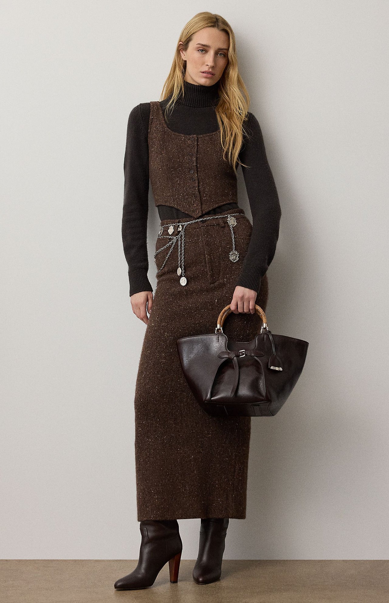 A woman wears a brown tweed vest and a matching RALPH LAUREN COLLECTION Tweed Ankle Pencil Skirt over a black turtleneck, accessorized with a layered silver chain belt and carrying a dark leather handbag with bamboo handles.