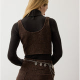 A woman with long blonde hair faces away, wearing a dark turtleneck under the RALPH LAUREN COLLECTION Tweed Scoop Neck Vest Top in a brown wool cashmere blend, paired with a matching skirt and a silver chain belt with charms.