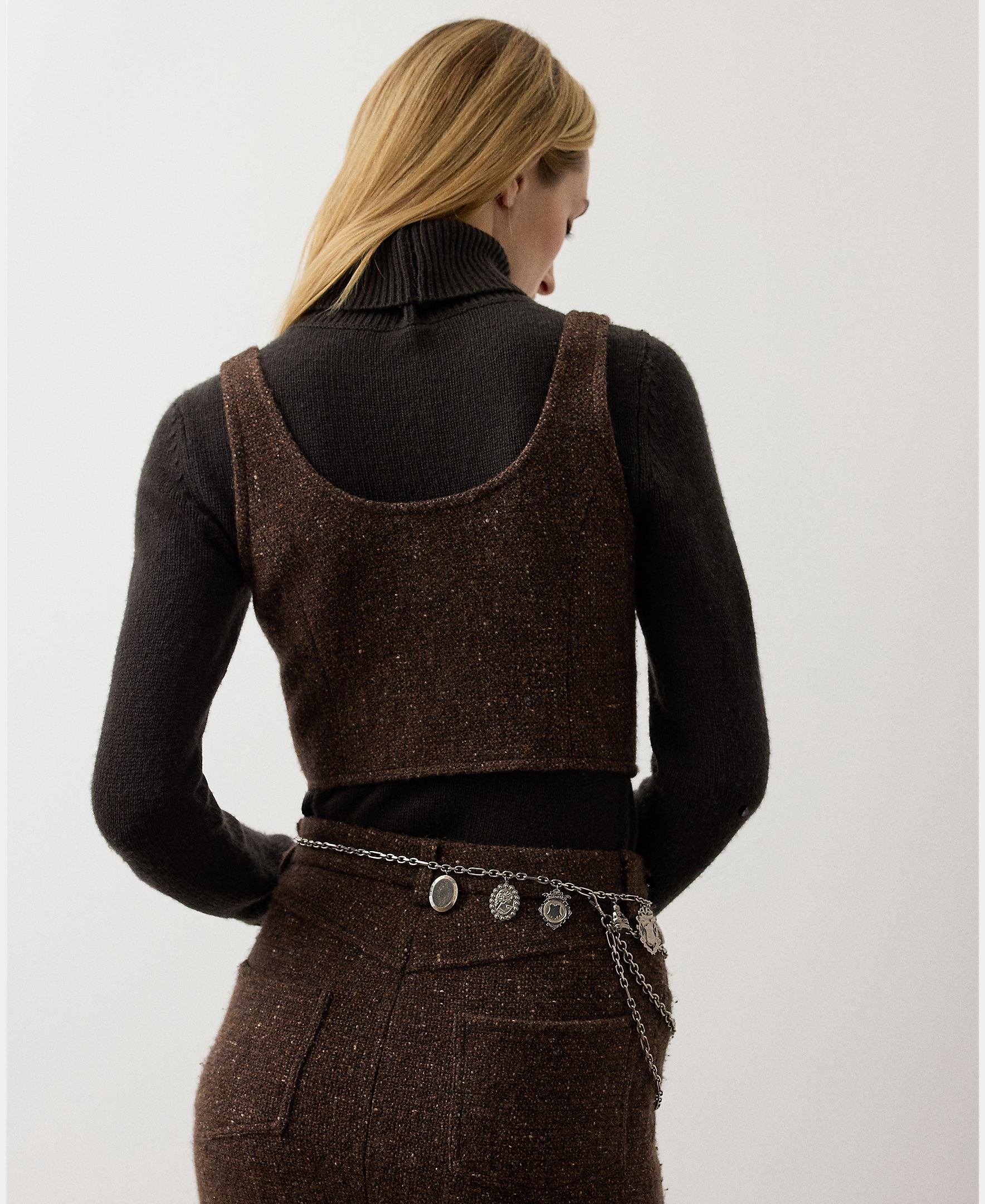 A woman with long blonde hair faces away, wearing a dark turtleneck under the RALPH LAUREN COLLECTION Tweed Scoop Neck Vest Top in a brown wool cashmere blend, paired with a matching skirt and a silver chain belt with charms.