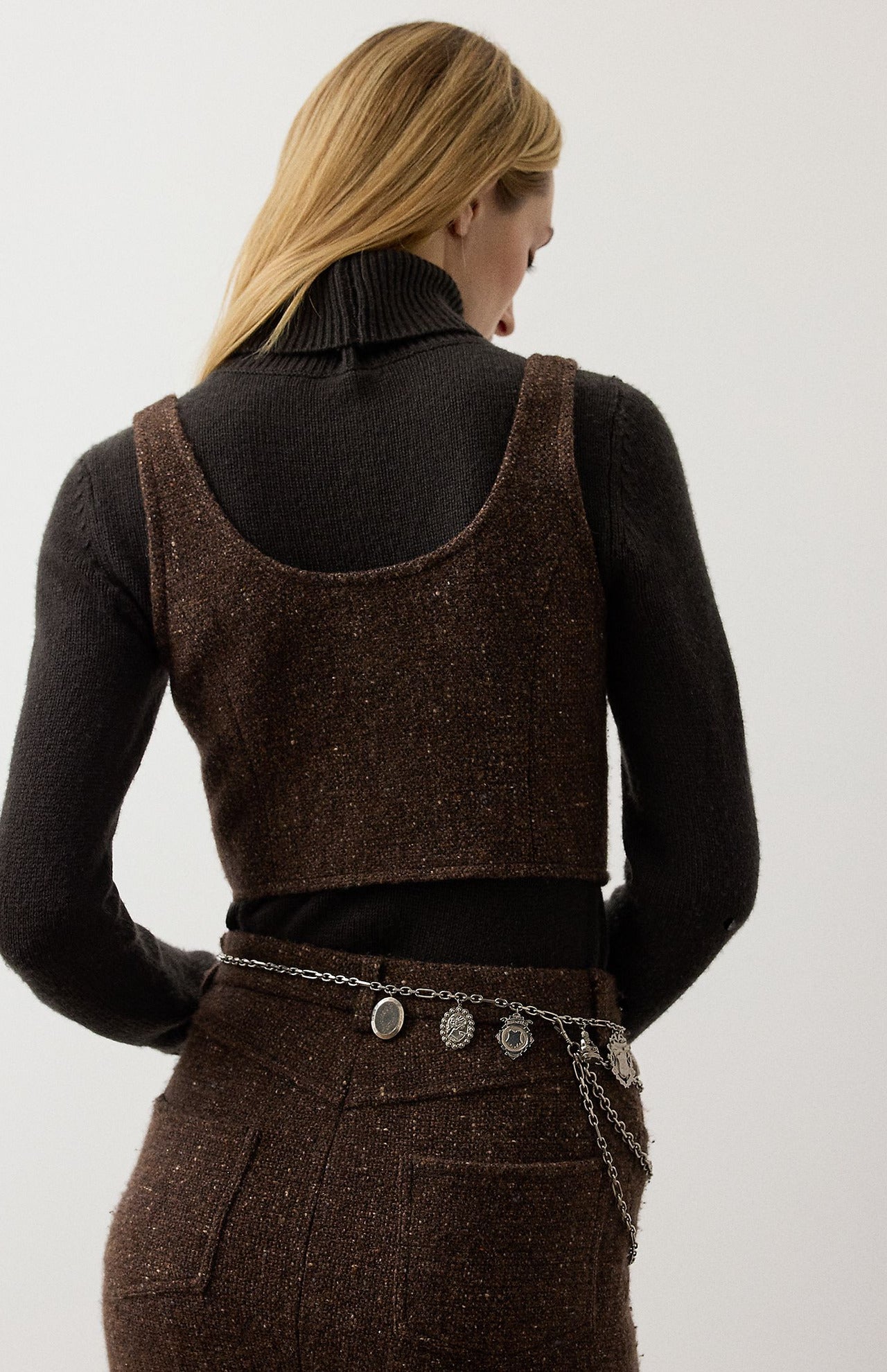A woman wears a brown Tweed Scoop Neck Vest Top over a dark brown turtleneck sweater with a matching tweed skirt and a dangling charm chain belt.