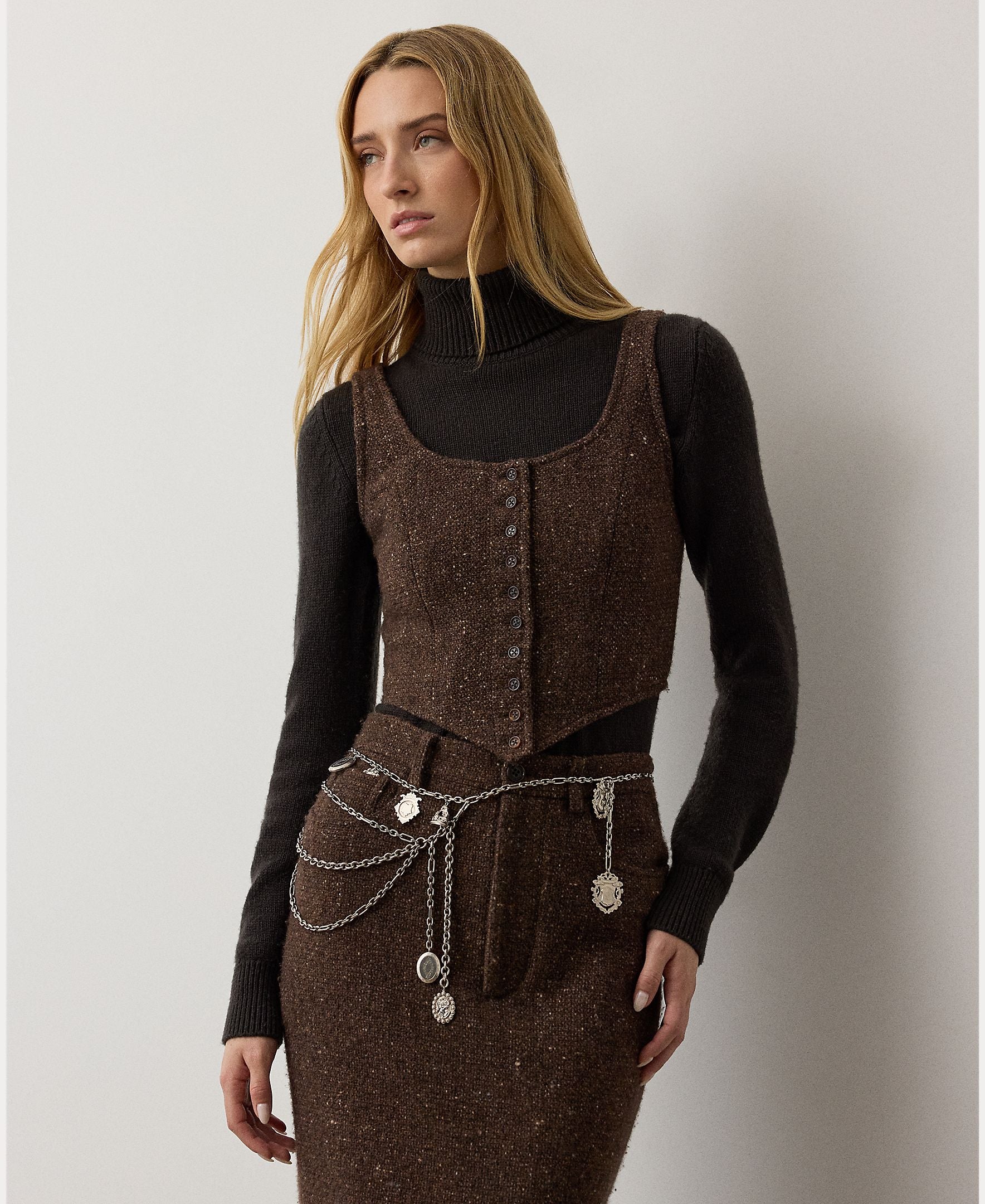 A woman with long blonde hair wears the RALPH LAUREN COLLECTION Tweed Scoop Neck Vest Top in dark brown wool cashmere blend, paired with a matching skirt, a black turtleneck, and a layered silver chain belt with pendants, standing against a plain light background.