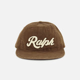 A brown cotton twill RALPH LAUREN POLO baseball cap featuring “Ralph” embroidered in large cream-colored script on the front, accented with classic logo embroidery and finished with an adjustable leather strap at the back.