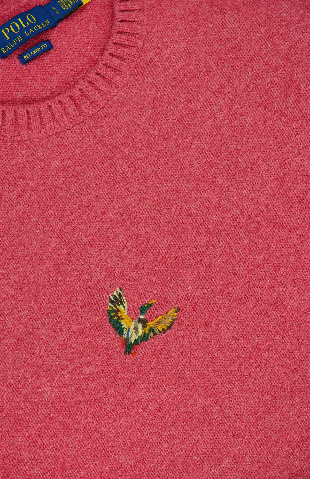 A close-up of the pink Ducks Fly Crewneck Sweater by RALPH LAUREN POLO, featuring a colorful embroidered bird logo and part of the blue and yellow brand tag visible at the neckline.