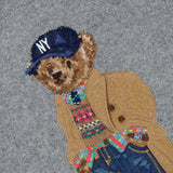 The Novelty Heritage Bear Crewneck Sweater by RALPH LAUREN POLO is a gray sweater featuring an embroidered bear wearing a blue NY cap, tan coat, colorful Fair Isle pullover, jeans, and sneakers. The Polo by Ralph Lauren label is visible at the neckline.