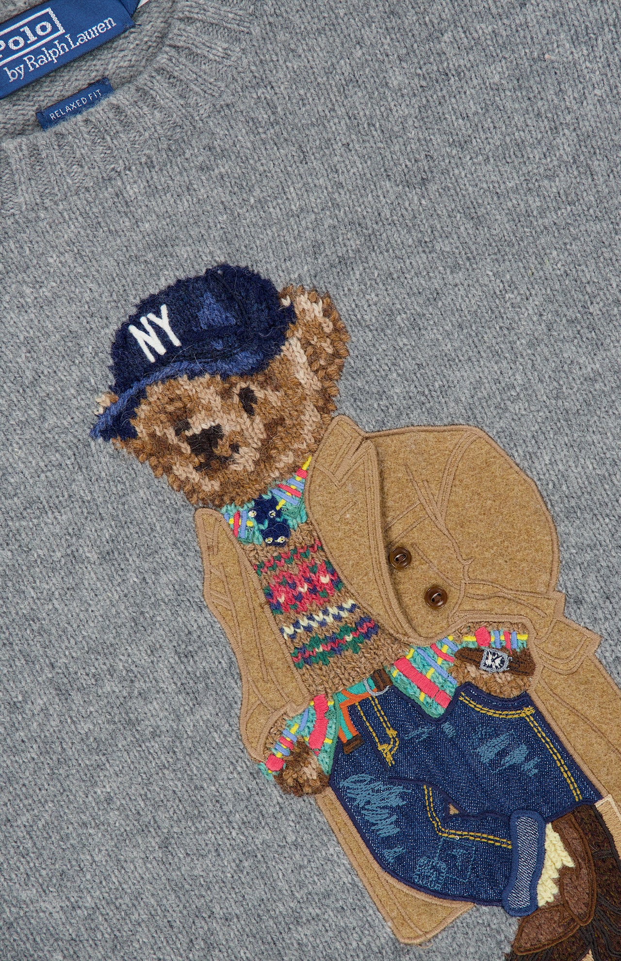 The Novelty Heritage Bear Crewneck Sweater by RALPH LAUREN POLO is a gray sweater featuring an embroidered bear wearing a blue NY cap, tan coat, colorful Fair Isle pullover, jeans, and sneakers. The Polo by Ralph Lauren label is visible at the neckline.