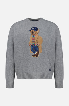 The Novelty Heritage Bear Crewneck Sweater by RALPH LAUREN POLO features a gray knit design with an embroidered bear dressed in a tan coat, blue jeans, colorful sweater, brown shoes, and a navy NY cap. Signature RL initials are stitched near the right side of the bottom hem, offering a classic finish inspired by timeless wool-cashmere sweaters.