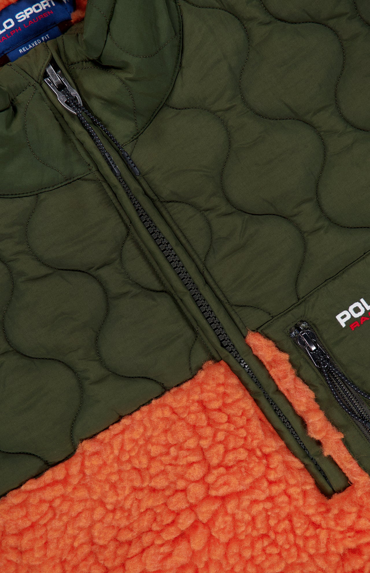 Close-up of the Shaggy 1/2 Zip Polo Sport Mockneck Sweater by RALPH LAUREN POLO in olive green, featuring black zippers and laid over bright orange fleece. The signature Polo Sport logo appears near the zipper, completing this bold color-blocked half-zip style.