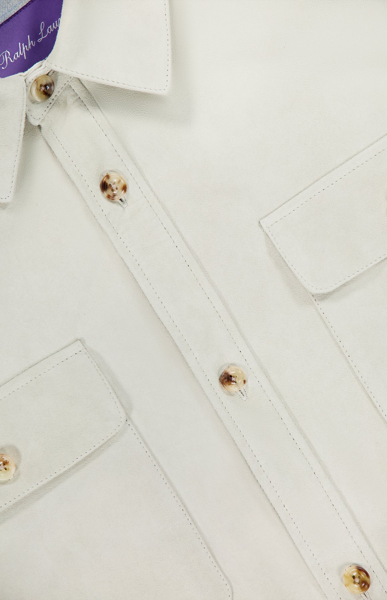 Close-up of the Calloway Suede Knit Overshirt by RALPH LAUREN PURPLE LABEL, featuring a white button-up design with two chest pockets, gold-tone buttons, and visible stitching. The edge of the signature purple label is visible inside the collar, showcasing Ralph Lauren Purple Label’s renowned sophistication and dedication to Italian craftsmanship.