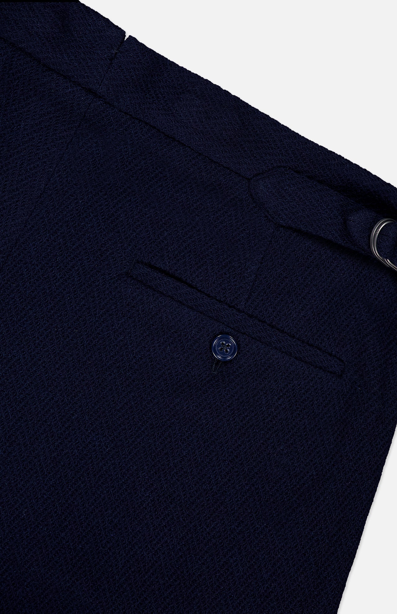 Close-up of the Cashmere Herringbone Pleated Tennis Short by RALPH LAUREN PURPLE LABEL, featuring navy blue textured fabric with a buttoned trouser pocket and a metal ring detail on the waistband, evoking classic Italian tailoring, set against a white background.